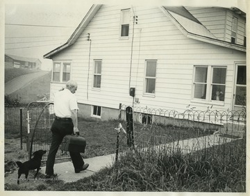 "Doc Harriman" carries a case and is followed by a small black dog through the fence. 