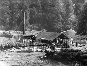 A group of men and women balance on large logs. Behind them are stacks of lumber. In the center of the photograph is saw mill, with a pipe that leads out into the forest and has created a mountain of sawdust.