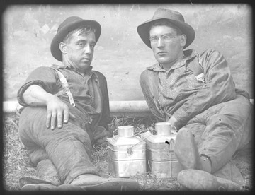 Two unidentified men lean against a tree branch with their lunch pails in between them. 