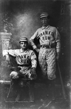 Two young men pose in their team uniforms. The player to the right is holding a baseball bat. 