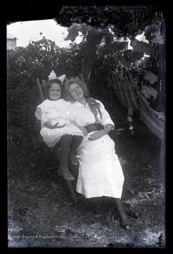 Two unidentified girls lean back in a lawn chair.  A hammock is visible to the right.