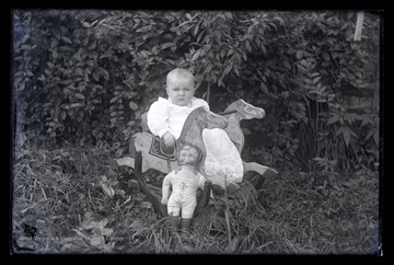 An unidentified infant is pictured sitting inside of a rocking horse. Beside the child is a doll. 