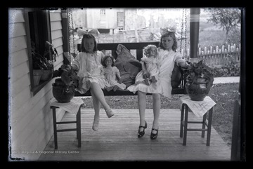 Two young girls sit on a porch swing accompanied with their dolls. 