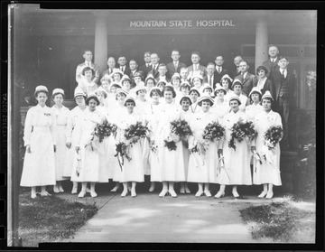 The nurses in the forefront hold flower bouquets. Behind them, physicians and staff are dressed in suits. 