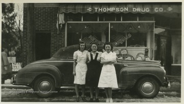 Three unidentified women pose beside a car parked in front of Thompson Drug Company. 