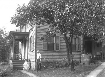 A man stands in a yard beside a house while a woman and child sit on the porch steps. Subjects unidentified. 