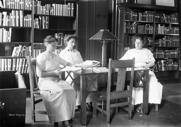 Three unidentified women read materials stacked on the table they are sitting at. The library was originally called the Hannah Fox Memorial Library. 