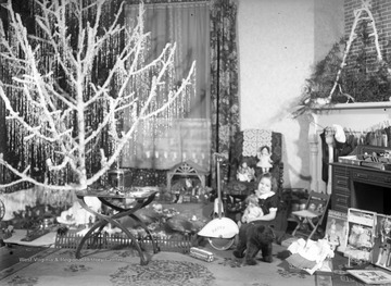 Young Patty Hoffman sits on the living room floor, surrounded by new toys. On the left of the photograph is a decorated Christmas tree. In the background to the right, several stocking hang from the fireplace. 