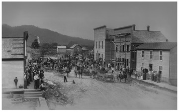 A mob returns to Parsons, W. Va. from the former seat of Tucker County, St. George.  The mob had taken records from the old courthouse by force.The view is from the corner of Main Street and Second Street, looking northwest down Main Street toward Alum Hill in the distance, which slopes downward to the right.There are two large buildings on the right.  The first one is under construction, and has scaffolding on it.  It was still standing in 2017 and houses the McClain Printing Co.  The second building, which is a little taller, is completed.  This building becomes the temporary courthouse, and the county records and furniture are put into it.  This building was torn down in 2007.