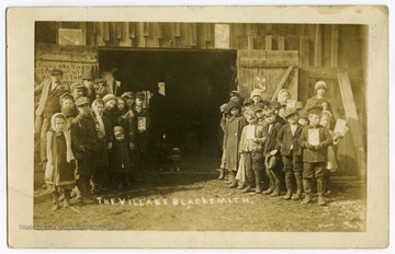 A group of children and some adults pose at the door of the blacksmith's shop at the south end of High Street.