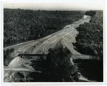 Text on the back reads, "The above aerial photo from the Department of Highways shows the area of Corridor "E" which includes the Cooper's Rock interchange in Cooper's Rock State Forest. The Harry Miller Excavating Company of Suffield, Ohio, is contractor for the work. Some 23 miles of construction currently is under way on the corridor in Monongalia and Preston counties at a cost of $50 million. The construction extends from near the western boundary of the forest all the way to the Maryland State Line, and also includes a three-mile segment which connects with Interstate 79 at Morgantown. Corridor "E" will be a four-lane highway from I-79 to the Maryland line for a total length of 31 miles."