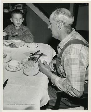 "Bud McDonald, right, unemployed, and Richard Love, boy belonging to another family in the community, eat a dinner comprised completely of agricultural commodities made available under the stepped-up food distribution program emphasized by President Kennedy. Various members of community were assembled to illustrate use of distributed foods. Ethel, W. Va."