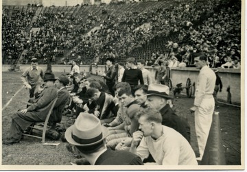 West Virginia University Football team on their benches, accompanied by coaches, cheerleaders and the Mountaineer mascot. The band can be seen in the stands behind them.